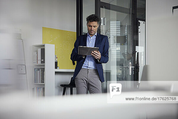 Businessman using tablet PC leaning on glass door at work place
