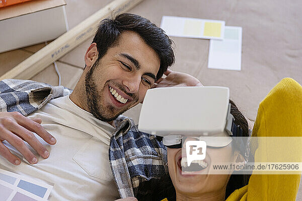 Glückliche Frau mit VR-Brille liegt neben ihrem Freund bei Renovierungsarbeiten zu Hause
