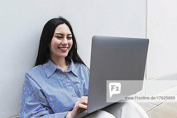 Smiling businesswoman using laptop sitting in front of white wall