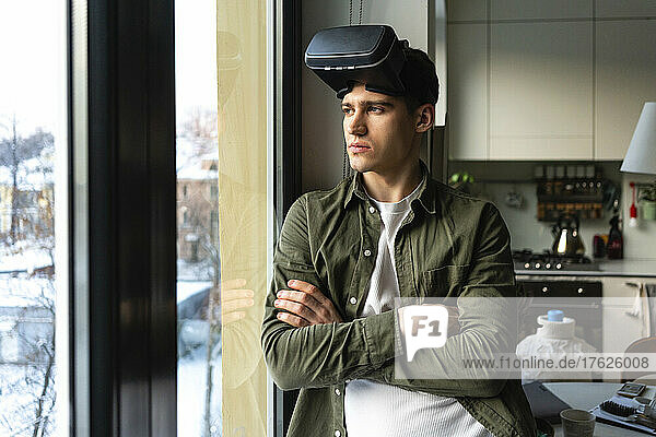Junger Mann mit Virtual-Reality-Simulator steht mit verschränkten Armen und blickt durch das Fenster