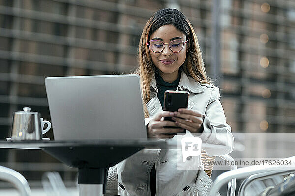 Smiling woman using smart phone sitting at sidewalk cafe