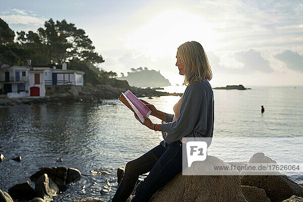 Frau liest Buch und sitzt auf einem Felsen am Strand