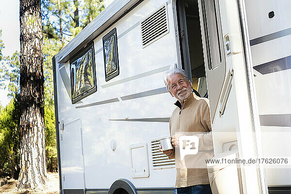 Smiling senior man with coffee cup standing at door of motor home