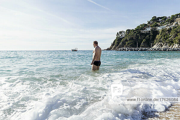 Mann mit nacktem Oberkörper steht am Strand im Meer