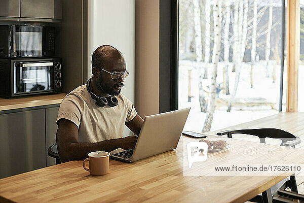 Man with headphones using laptop on table in kitchen at home