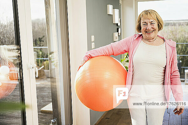 Happy woman with fitness ball standing at glass window at home
