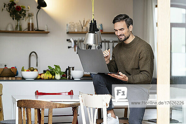 Smiling young man using laptop siting on table in kitchen at home