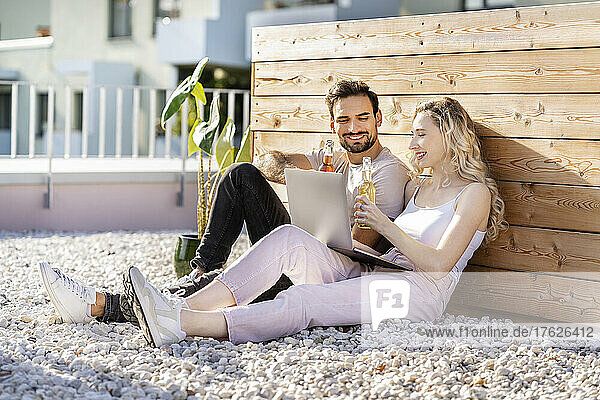 Smiling young couple toasting beer sitting with laptop on sunny day