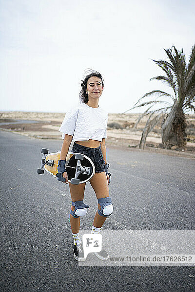 Lächelnde junge Frau mit Skateboard  die auf der Straße läuft