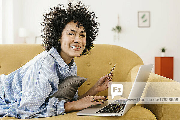 Happy young woman with credit card and laptop lying on sofa at home