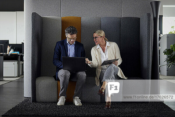 Businesswoman discussing with colleague using laptop sitting on sofa in office