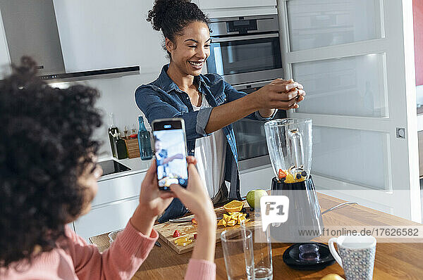 Woman photographing friend making juice in kitchen at home