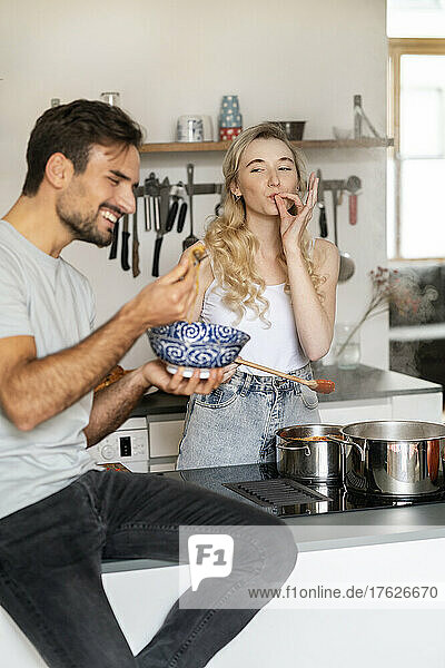 Woman gesturing ok sign and looking at boyfriend eating spaghetti in kitchen