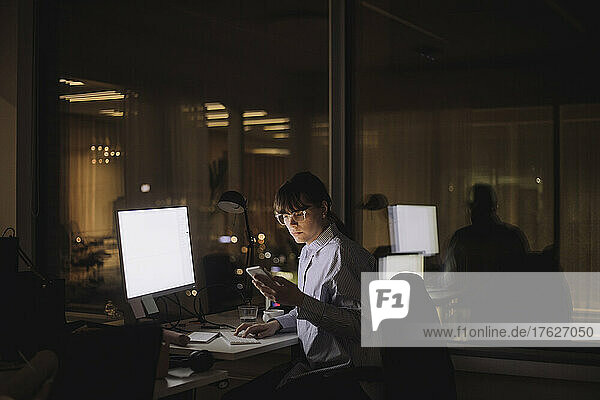 Businesswoman text messaging on mobile phone sitting in office at night