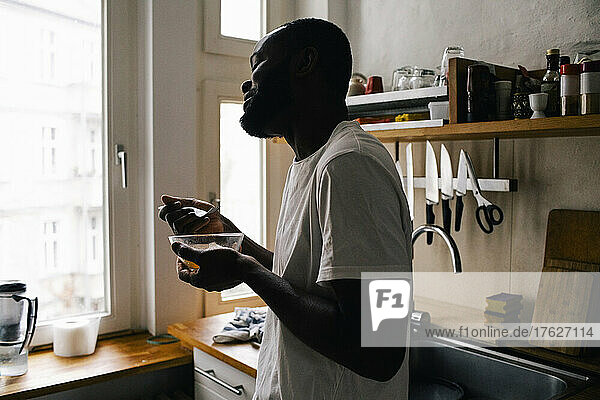 Seitenansicht eines lächelnden Mannes  der eine Schüssel und eine Gabel hält und in der Küche zu Hause isst
