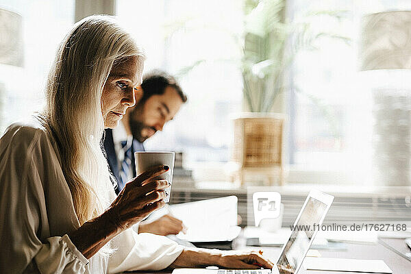 Mature businesswoman holding coffee mug using laptop by businessman at desk in office
