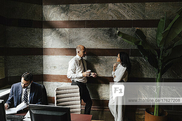 Geschäftskollegen diskutieren  während sie sich an die Wand in der Nähe eines Geschäftsmannes lehnen  der am Schreibtisch im Büro arbeitet