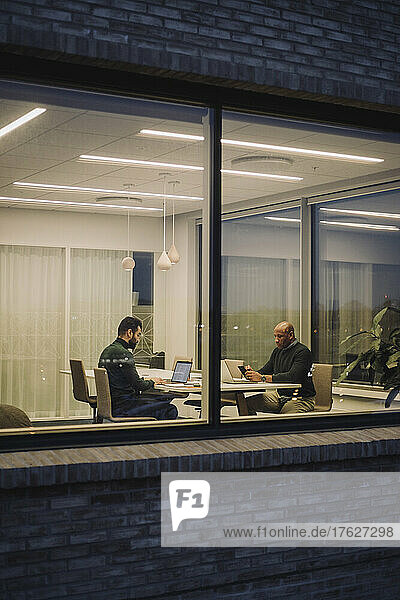 Geschäftsmann mit männlichem Kollegen  der spät im Büro arbeitet  gesehen durch ein Fenster bei Nacht