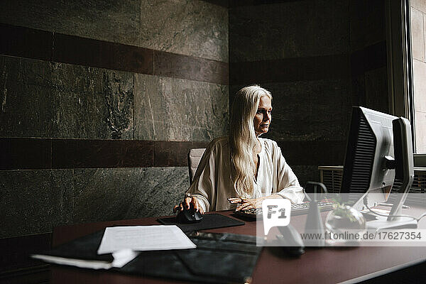 Selbstbewusste reife Geschäftsfrau mit langen weißen Haaren  die einen Computer am Schreibtisch im Büro benutzt