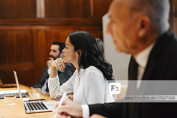 Geschäftsfrau mit Hand am Kinn sitzt inmitten männlicher Kollegen in einer Vorstandssitzung