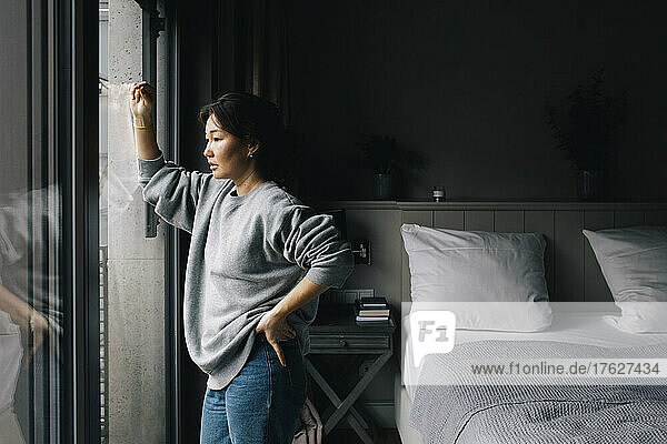 Nachdenkliche einsame Frau  die aus dem Fenster schaut  während sie mit der Hand auf der Hüfte im Schlafzimmer zu Hause steht