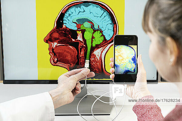 Close-up of hands holding a smartphone and connecting it to a brain image on a computer screen.