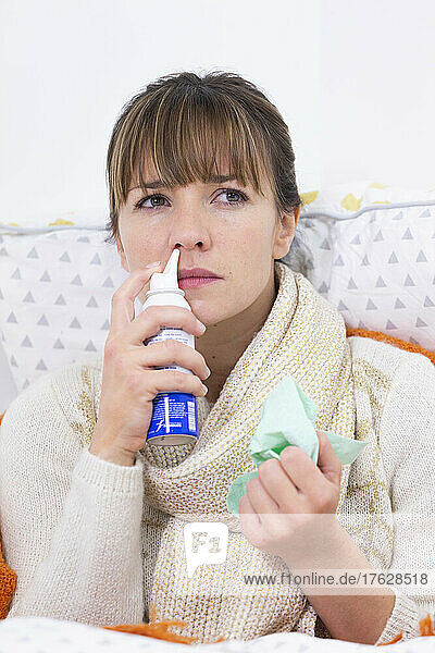 Young woman in bed suffering from a cold with nasal spray.