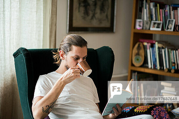 Man drinking coffee and reading book in armchair