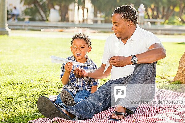 Glücklicher afroamerikanischer Vater und gemischtrassiger Sohn spielen mit Papierflugzeugen im Park