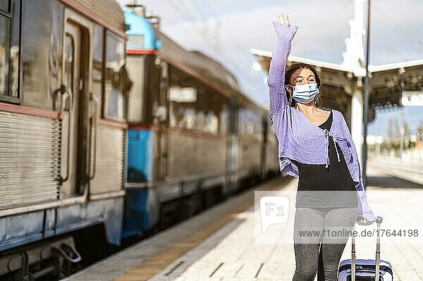 A young woman in mask and with luggage greetings somebody at the platform of train station