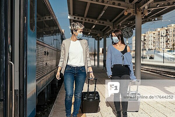 Two happy female friends in protective masks walking and chatting on the platform at train station