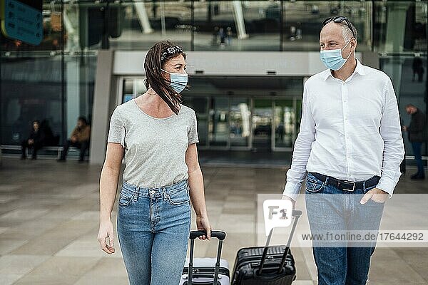 A couple of friends in protective masks chatting while leaving the airport with their luggage