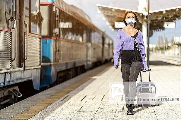 A young woman in a mask and with luggage walking on the platform of a train station at her destination