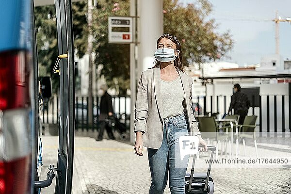 A young woman wearing protective maedical mask boarding the bus with a luggage