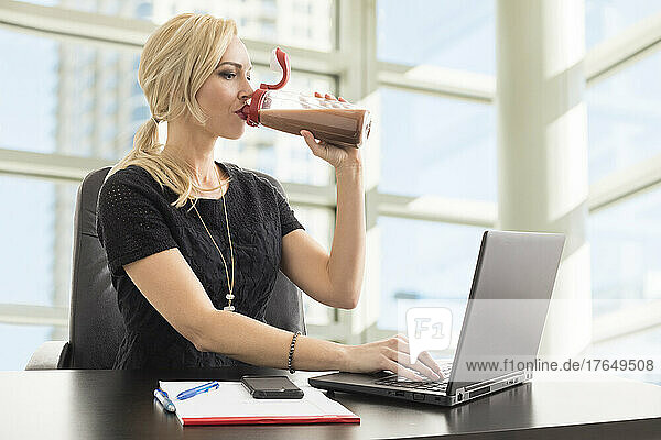 Businesswoman using laptop and drinking protein shake in office
