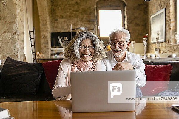 Happy woman using laptop sitting with man on sofa at boutique hotel