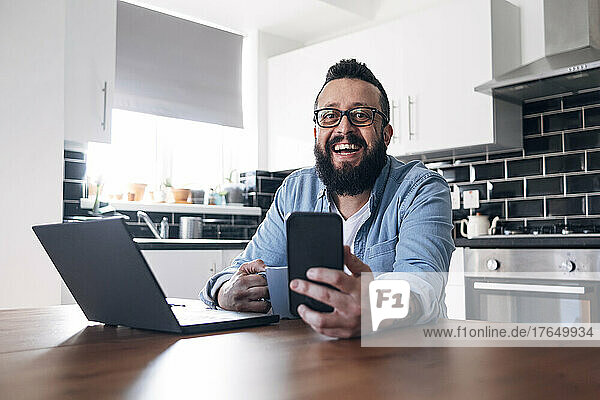 Happy freelancer with laptop and smart phone sitting in kitchen at home