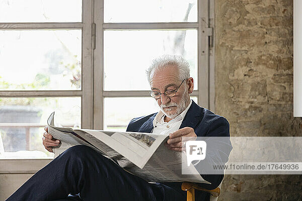 Senior man reading newspaper sitting in front of window at home
