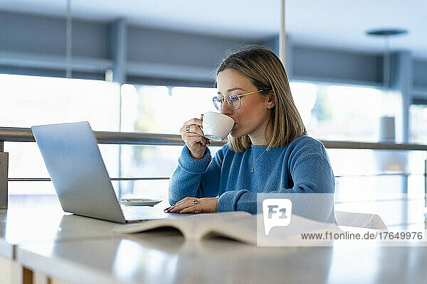 Young woman drinking coffee sitting with laptop and book at table