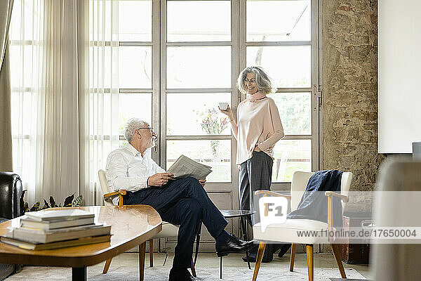 Smiling senior woman holding coffee cup talking with man in living room at home