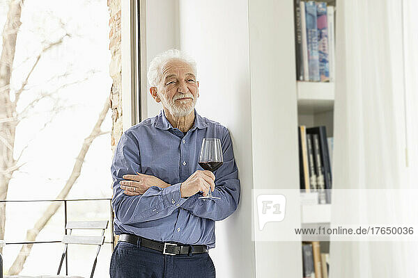 Senior man holding wineglass leaning on wall in front of window at home