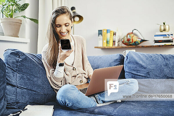 Happy freelancer sitting with laptop talking on smart phone through speaker in living room at home