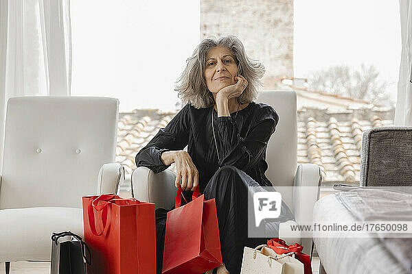 Smiling senior woman with hand on chin and shopping bags sitting on armchair at boutique hotel