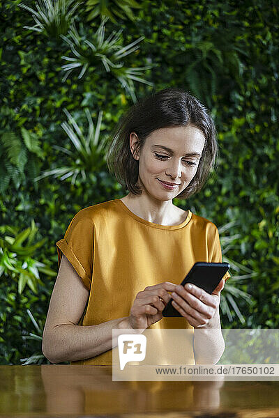Smiling woman using smart phone at wall with plants