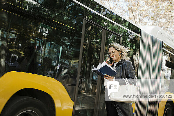 Smiling woman talking on smart phone and reading diary by bus
