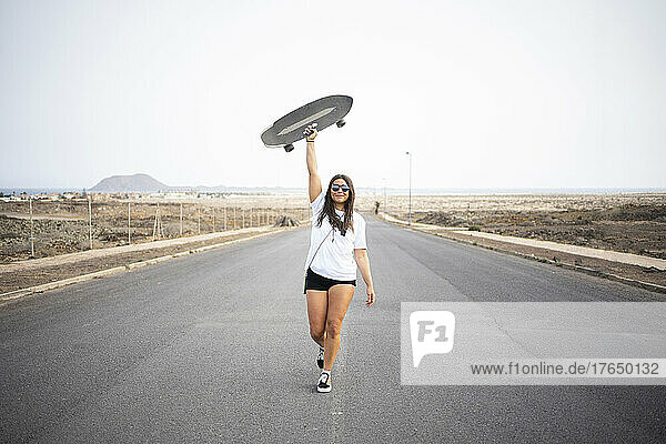 Smiling woman holding skateboard aloft on road