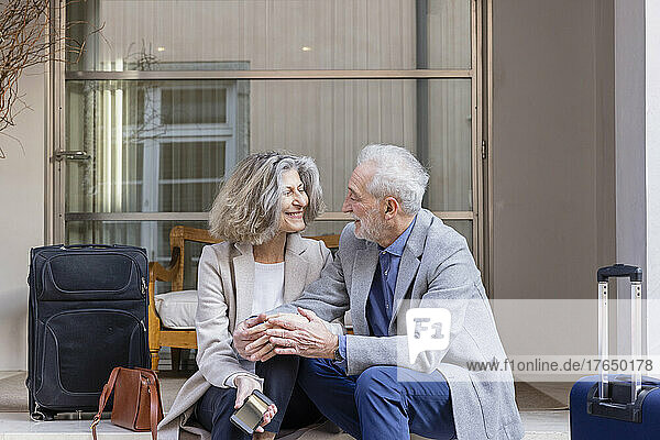 Happy senior couple with luggage sitting outside boutique hotel