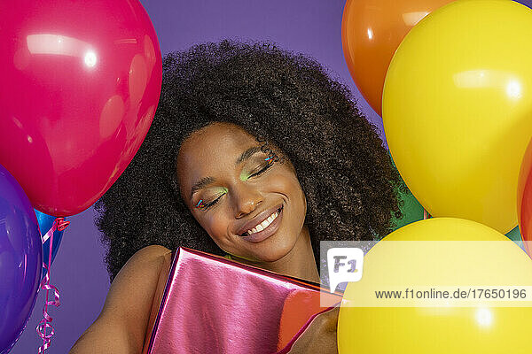 Fröhliche junge Frau mit geschlossenen Augen hält eine Geschenkbox in der Hand und steht inmitten bunter Luftballons vor violettem Hintergrund