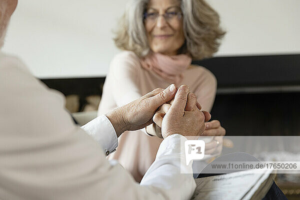 Älterer Mann hält Frau an der Hand  die zu Hause auf einem Stuhl sitzt