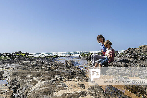 Happy mother and daughter sitting on rock at sunny beach